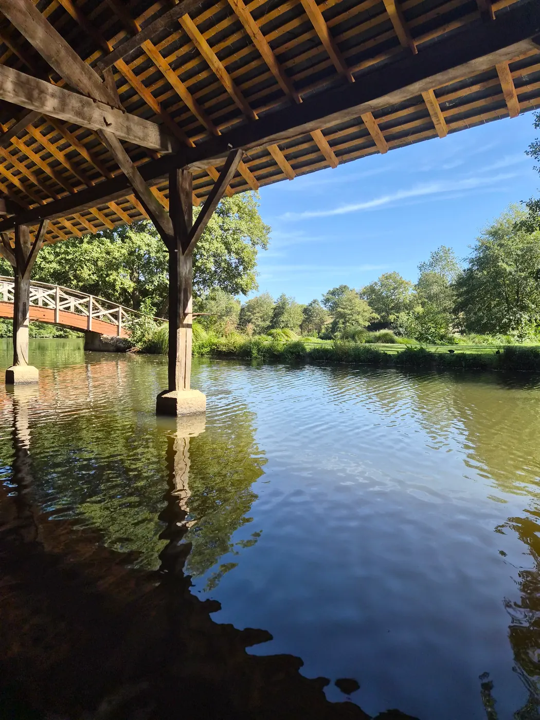 Sous la charpente du lavoir : les piles de bois se reflètent dans l'eau du plan d'eau.
